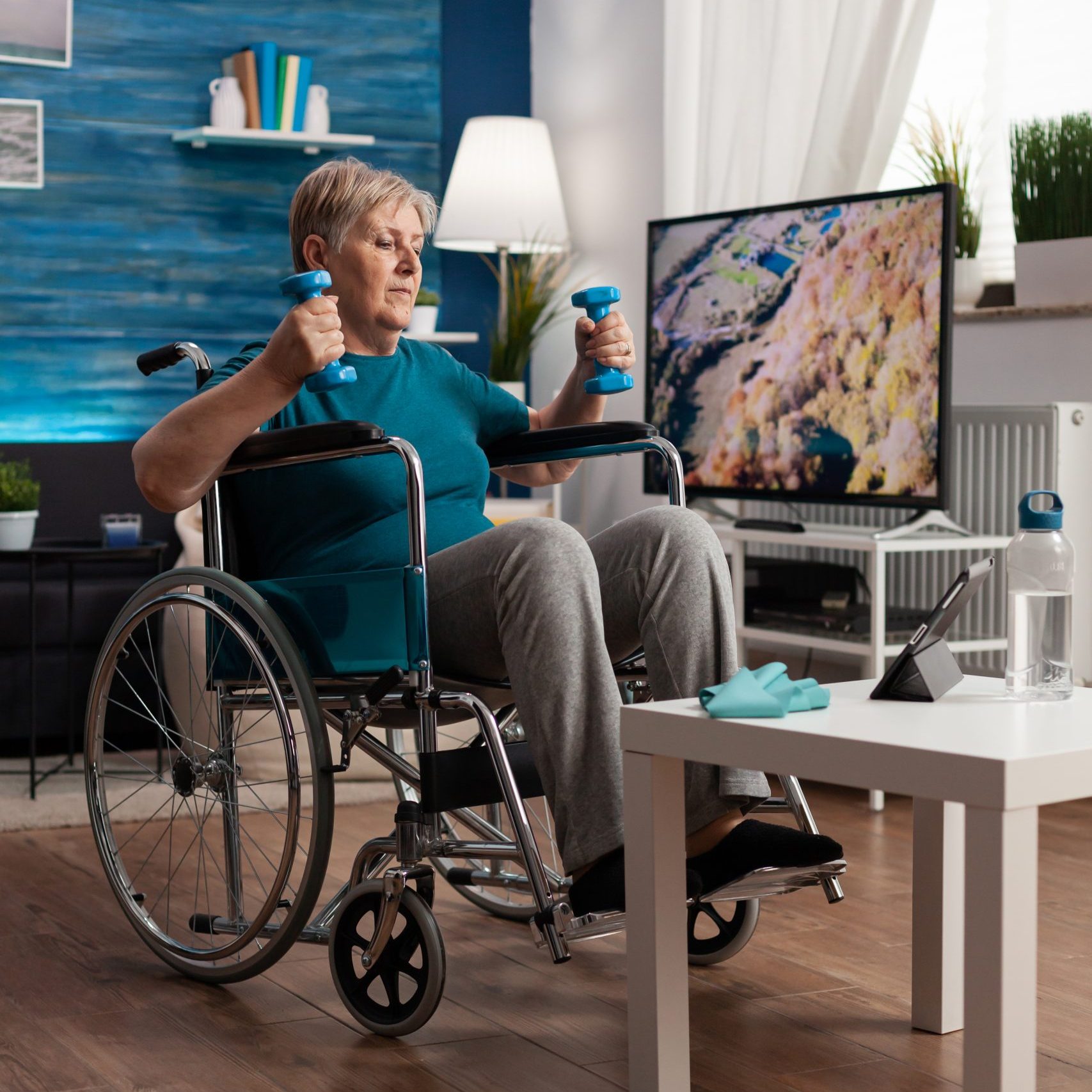 Handicapped senior woman in wheelchair stretching arms muscles exercising body resistance using workout dumbbells in living room. Handicapped pensioner looking at lifestyle aerobics video on tablet